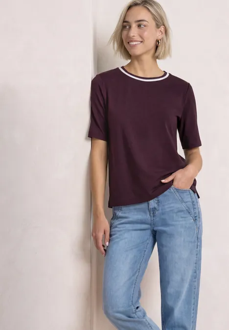 Eine lächelnde Frau in lila T-Shirt mit weißem Kragen und blauen Jeans lehnt an einer Wand.