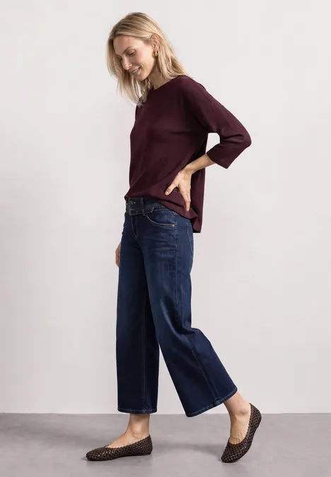Frau in bordeauxrotem Oberteil, weiten blauen Jeans und braunen flachen Schuhen.
