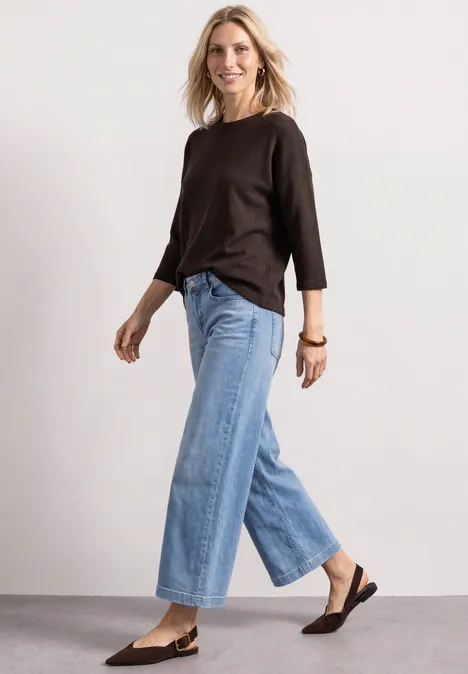 Frau in braunem Oberteil, weitem Jeans und braunen Loafers vor neutralem Hintergrund, geht lächelnd.