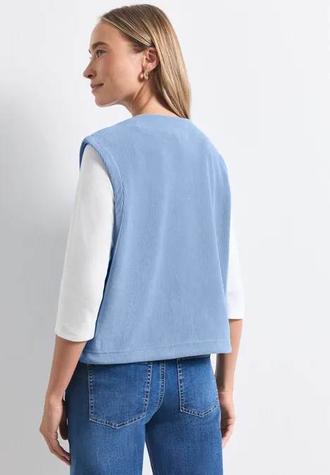 Eine Frau von hinten in hellblauer Weste über weißem Shirt und Jeans vor hellem Hintergrund