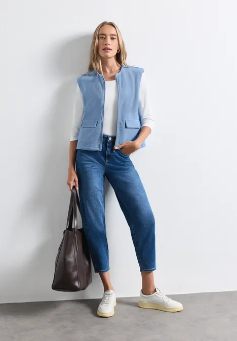 Frau in weißer Bluse, hellblauer Weste, blauen Jeans, weißen Sneakern und mit brauner Tasche.