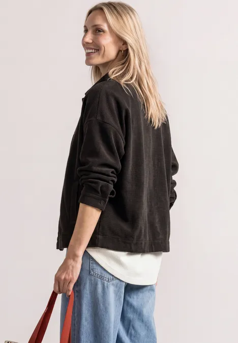 Femme souriante regarde par-dessus son épaule, porte veste noire, chemisier blanc, jean bleu et sac rouge.