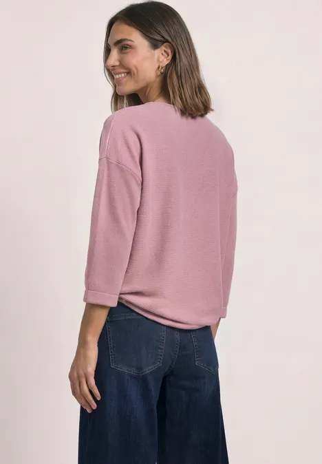 Eine Frau mit rosa Dreiviertelarmshirt und weiten Jeanshosen schaut über die Schulter und lächelt.