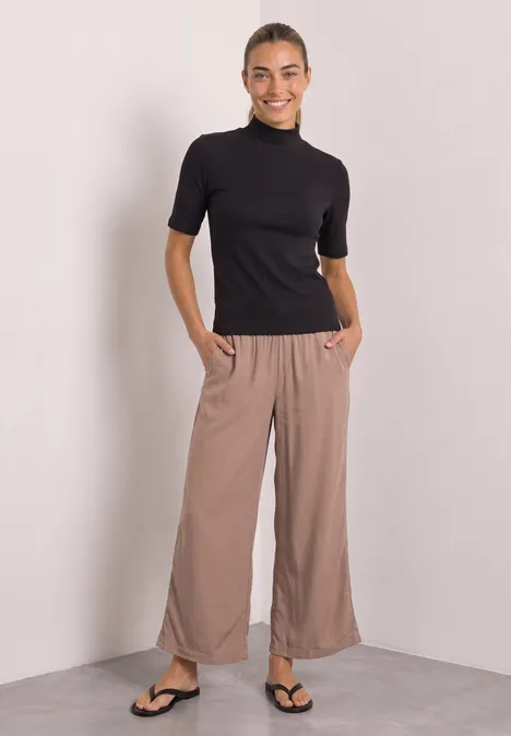 Frau in schwarzem Kurzarm-Rollkragenpullover, beigen, weiten Hosen und Flip-Flops lächelt entspannt.