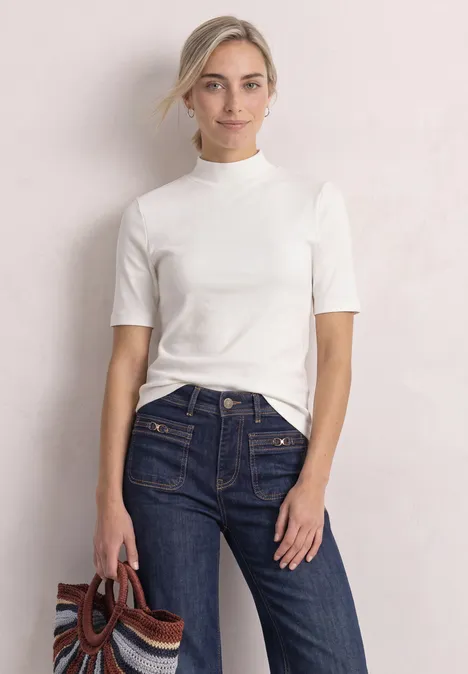 Shirt met col in ribbelpatroon off white Een vrouw met los teruggebonden haar draagt een wit shirt en donkere jeans voor een lichte achtergrond.