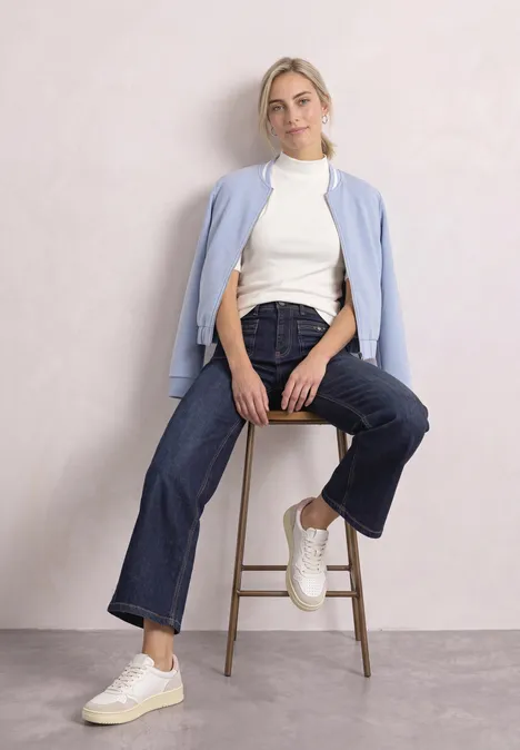 Shirt met col in ribbelpatroon off white Vrouw zit op kruk voor lichte muur, draagt witte sneakers, wijde jeans, witte coltrui en lichtblauwe cardigan.
