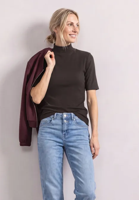 Frau in dunklem Kurzarmshirt, Jeans und weinroter Jacke über der Schulter, lächelnd vor neutralem Hintergrund.