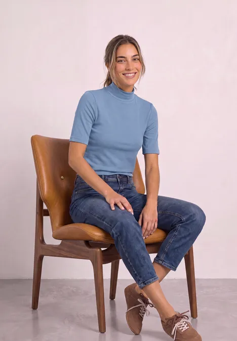 Shirt met col in ribbelpatroon northern blue Junge Frau in hellblauem T-Shirt und Jeans sitzt lächelnd auf einem Holzstuhl.