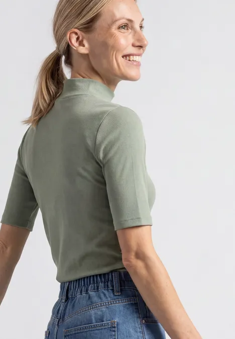 Eine Frau in einem olivgrünen Kurzarmshirt und Jeans schaut seitlich und lächelt.