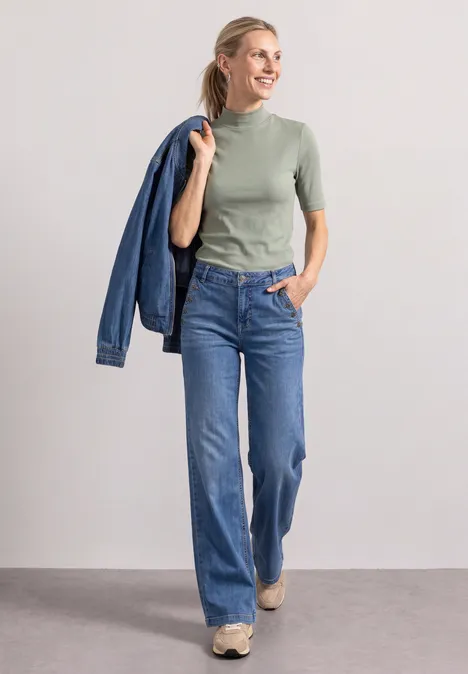 Eine Frau in grüner Kurzarmbluse und blauen Jeans, lächelnd und eine Jeansjacke über der Schulter tragend.