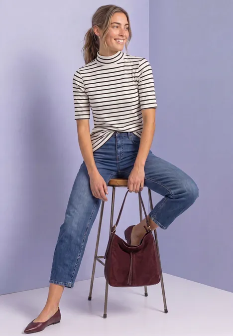 Junge Frau in gestreiftem Shirt und Jeans sitzt auf einem Hocker, hält eine weinrote Handtasche vor lila Hintergrund.