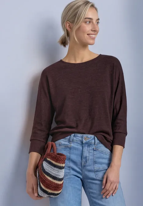 Batwing-Shirt in gemêleerde look rustic rouge mel. Vrouw in bruine trui en jeans met blond staartje houdt een kleurrijke gestreepte clutch vast en lacht.
