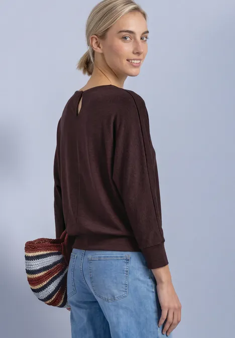 Batwing-Shirt in gemêleerde look rustic rouge mel. Vrouw in een bruine trui en spijkerbroek draagt een kleurrijke gestreepte tas en glimlacht.