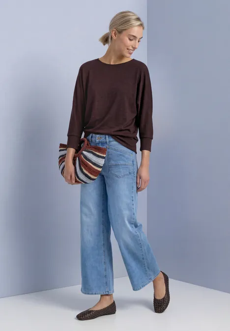 Batwing-Shirt in gemêleerde look rustic rouge mel. Vrouw met blond haar in knot, donkerrood trui, lichtblauwe wijde jeans, bruine ballerina’s en gestreepte tas voor effen achtergrond.