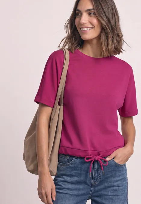 Frau in fuchsiafarbenem Oberteil mit Kordelbund, blaue Jeans und beiger Tasche, lächelnd.