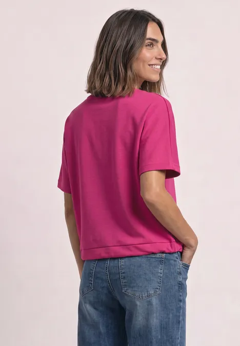 Seitlich lächelnde Frau in pinkem T-Shirt und Jeans, Hände in den Taschen.