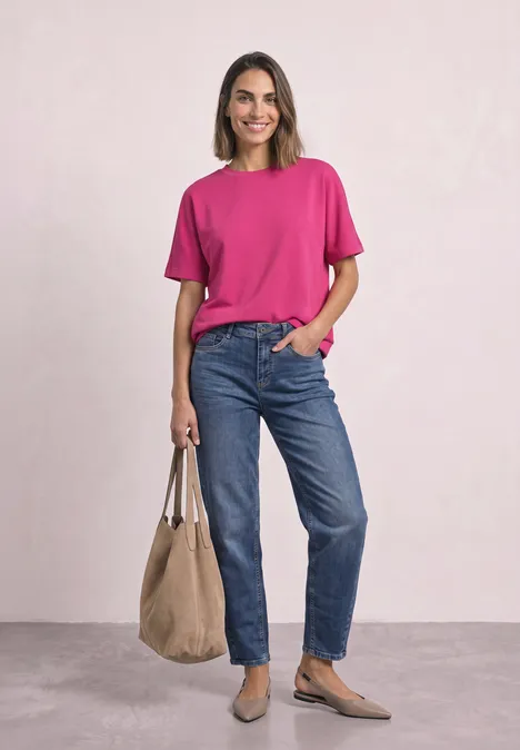 Lächelnde Frau in pinkem T-Shirt und blauen Jeans mit beiger Tasche vor hellem Hintergrund.