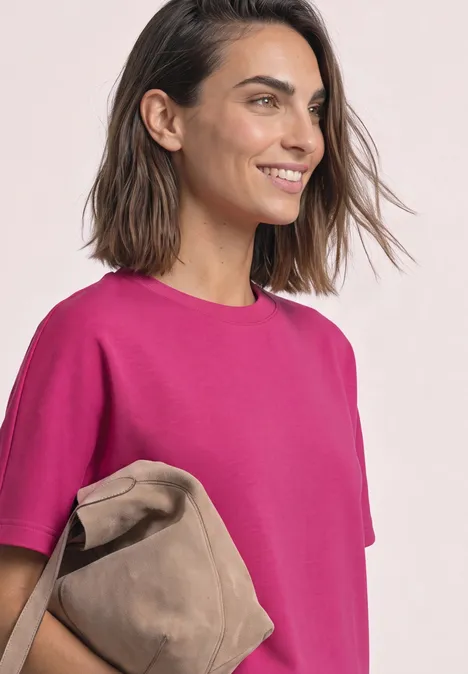 Frau lächelt leicht seitlich, trägt pinkes T-Shirt und hält eine beige Tasche.