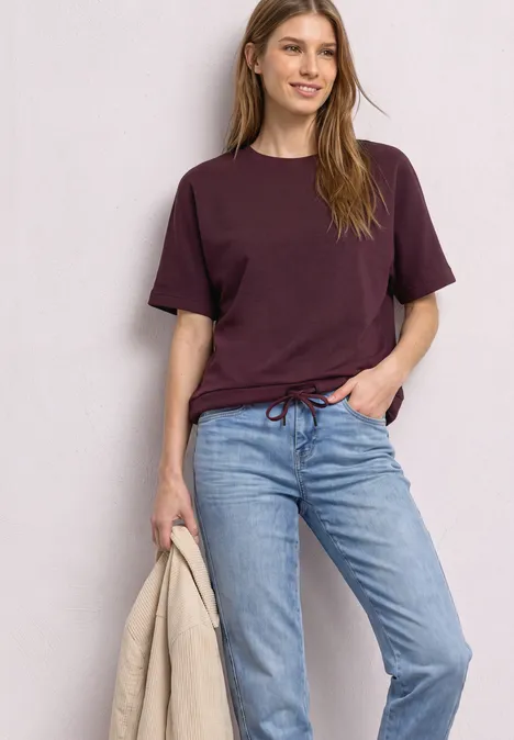 T-Shirt mit Rundhals im Silk-Look rustic rouge Eine lächelnde Frau trägt ein bordeauxrotes T-Shirt und Jeans, hält eine beige Jacke in der Hand.