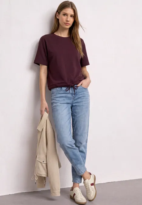 T-Shirt mit Rundhals im Silk-Look rustic rouge Junge Frau lehnt an weißer Wand, trägt weinrotes T-Shirt, hellblaue Jeans, weiße Sneaker und hält eine beige Jacke.