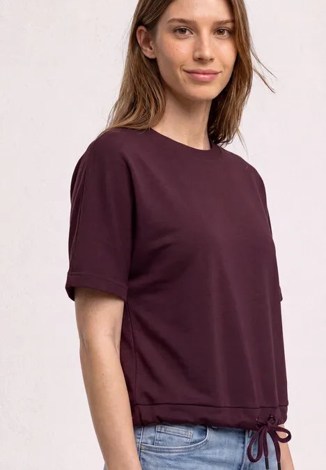 T-Shirt mit Rundhals im Silk-Look rustic rouge Eine junge Frau trägt ein kurzärmeliges, pflaumenfarbenes T-Shirt mit Tunnelzugsaum und blaue Jeans.