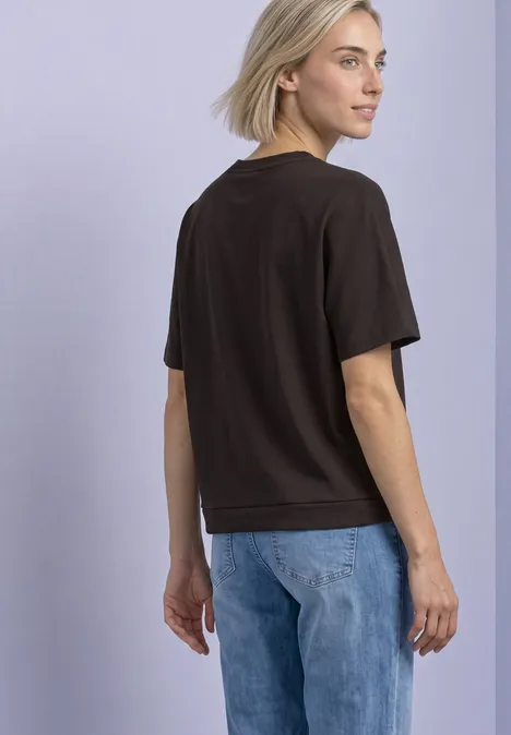 T-Shirt mit Rundhals im Silk-Look black coffee Blonde Frau in schwarzem T-Shirt und blauen Jeans, steht seitlich und blickt über die Schulter vor hellem Hintergrund.