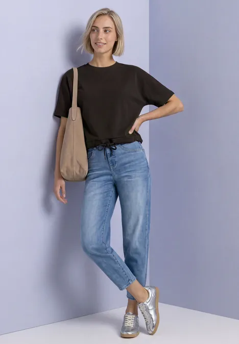 T-Shirt mit Rundhals im Silk-Look black coffee Frau in schwarzem T-Shirt, blauen Jeans und Sneakers, mit Stofftasche vor lila Hintergrund.