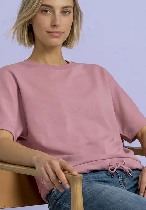 T-Shirt mit Rundhals im Silk-Look vintage blush Frau auf Stuhl in hellem Raum, trägt rosa T-Shirt mit Kordelzug und blaue Jeans.