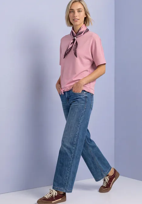 T-Shirt mit Rundhals im Silk-Look vintage blush Frau in rosa Shirt mit Halstuch, blauen Jeans und Sneakers vor hellviolettem Hintergrund.