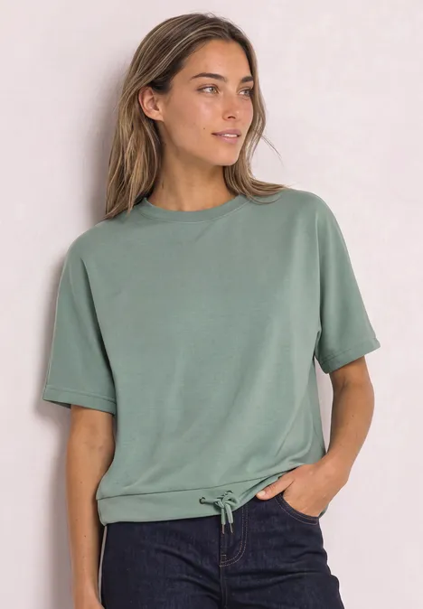 Frau trägt legeres grünes T-Shirt mit Tunnelzug und dunkler Hose vor hellem Hintergrund.