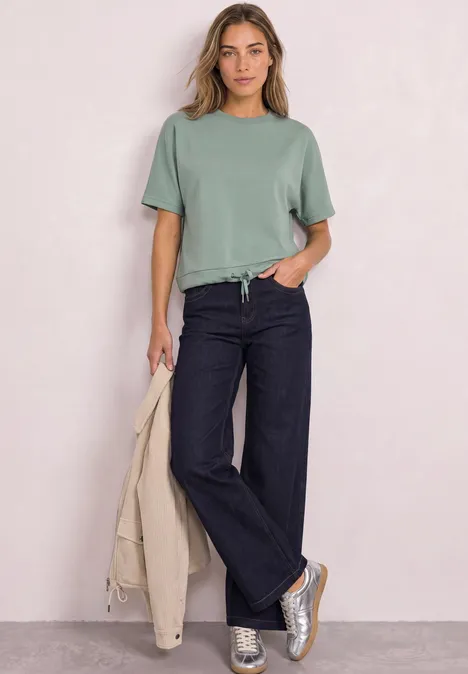 Frau in grünem T-Shirt, dunklen Schlagjeans und Sneakern, hält eine beige Jacke.
