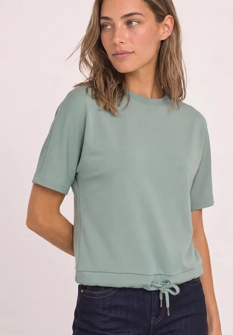 Eine junge Frau trägt ein salbeigrünes, kurzärmeliges Sweatshirt mit Tunnelzug am Saum und eine dunkle Jeans.