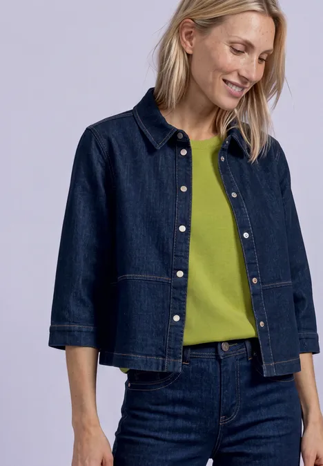 Une femme porte une veste en jean foncé, un jean et un t-shirt vert, souriant en regardant vers le bas.