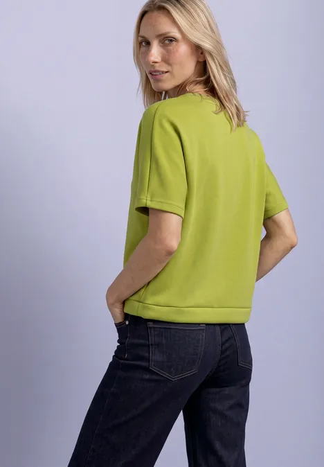 Femme en chemisette verte et jean foncé regarde par-dessus son épaule, mains dans les poches.