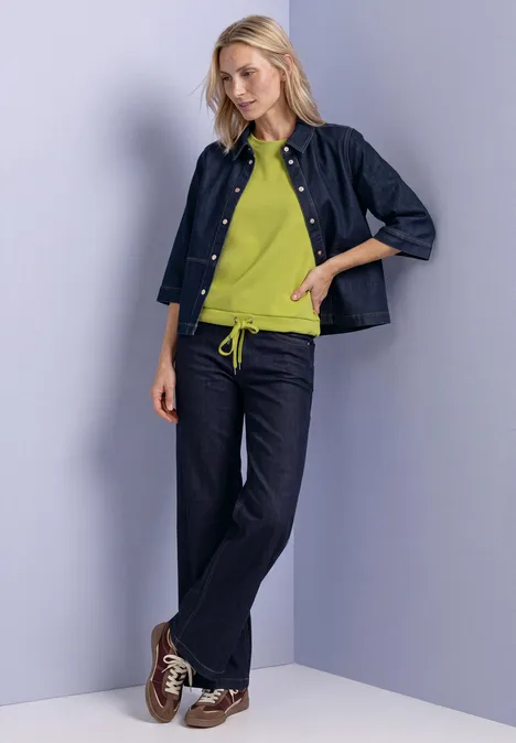 Femme en veste et jean denim, pull vert, appuyée de façon décontractée contre un mur.