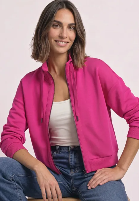 Une femme souriante en veste à capuche rose, débardeur blanc et jean sur fond clair.