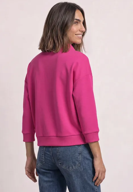 Femme de dos portant un sweat rose et un jean, tête légèrement tournée avec un sourire.