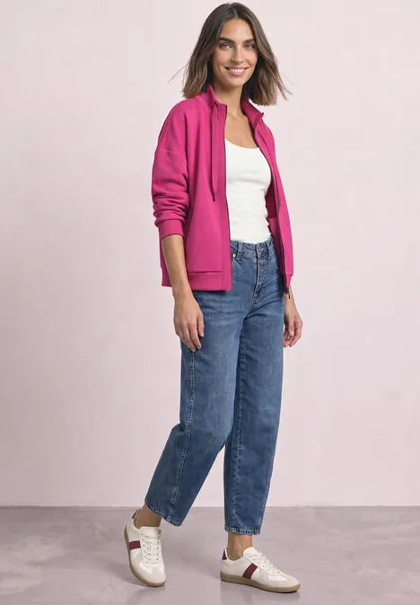 Femme souriante en sweat à capuche rose, top blanc, jean bleu et baskets blanches.