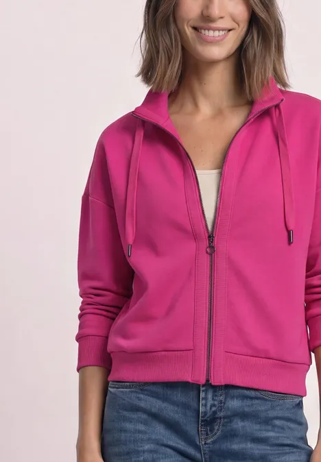 Une femme porte un sweat à capuche zippé rose vif et un jean bleu.