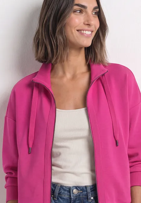 Une femme porte une veste sweat rose fuchsia à zip, débardeur blanc et sourit.