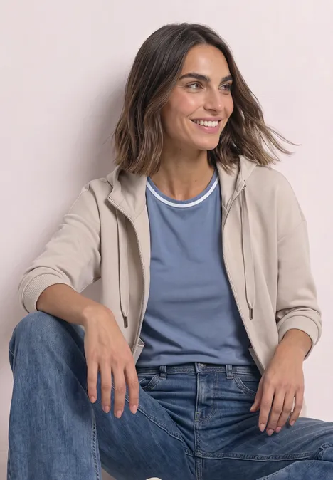 Jacke im Silk-Look mit Rippdetails clay sand Eine lächelnde Frau sitzt entspannt in Jeans, blauem T-Shirt und beiger Kapuzenjacke vor hellem Hintergrund.