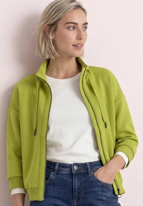 Femme aux cheveux blonds courts porte un sweat vert à zip, un t-shirt blanc et un jean bleu.