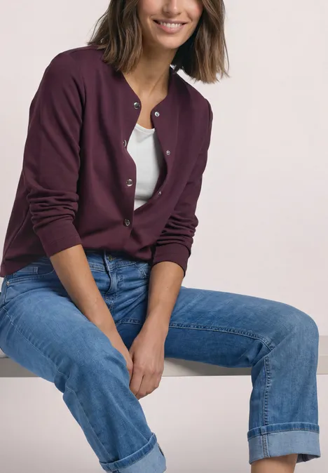 Strick-Look Jacke mit dekorativen Knöpfen rustic rouge Eine Frau sitzt lächelnd, trägt eine lila Strickjacke, weiße Bluse und blaue umgeschlagene Jeans.