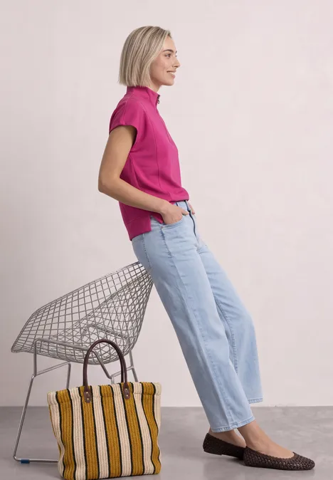 Mouwloos shirt met Silk-Look en ritssluiting magenta dream Frau in pinkem Shirt sitzt auf Drahtstuhl, trägt weite Jeans und flache Schuhe, neben ihr steht eine gestreifte Tasche.