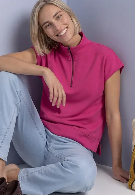 Mouwloos shirt met Silk-Look en ritssluiting magenta dream Eine lächelnde Frau sitzt auf dem Boden, lehnt an einer Wand, trägt pinkes Oberteil und helle Jeans.