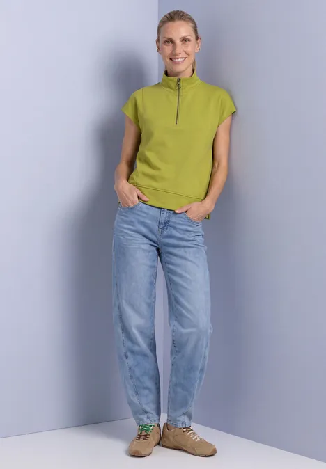 Mouwloos shirt met Silk-Look en ritssluiting apple lime Een vrouw in lichtblauwe spijkerbroek, groen kortärmelig shirt met rits en bruine schoenen voor een grijze achtergrond.