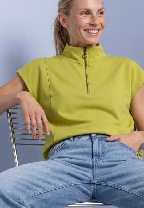 Mouwloos shirt met Silk-Look en ritssluiting apple lime Een glimlachende vrouw in groen topje en spijkerbroek zit ontspannen op een stoel.