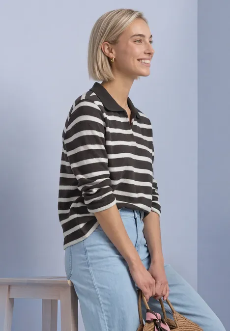 Polo met 3/4-mouwen en streepjespatroon black coffee Een glimlachende vrouw met kort blond haar, gestreepte trui en lichtblauwe spijkerbroek leunend opzij.