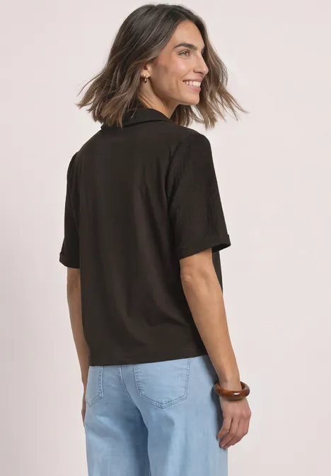 Shirt met verschillende texturen en een knoopsluiting black coffee Vrouw met kort bruin haar draagt een zwart poloshirt met korte mouwen en een lichte spijkerbroek, gezien van achteren.