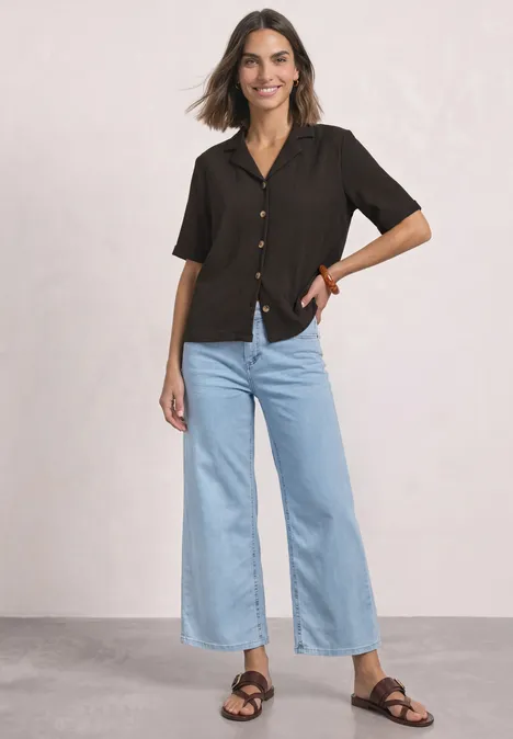 Shirt met verschillende texturen en een knoopsluiting black coffee Vrouw lacht in een zwarte kortemouw blouse, wijd lichtblauwe cropped jeans en zwarte sandalen.